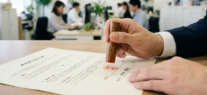 明るいオフィスで書類（業務委託契約書）に木目調の印鑑を押印するビジネスマンの手元のアップ